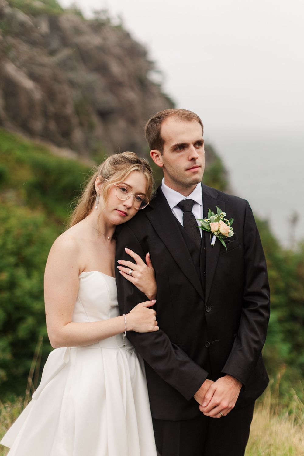 Bride in white strapless gown leans on groom in black suit with boutonniere, posing outdoors near rocky cliffs.