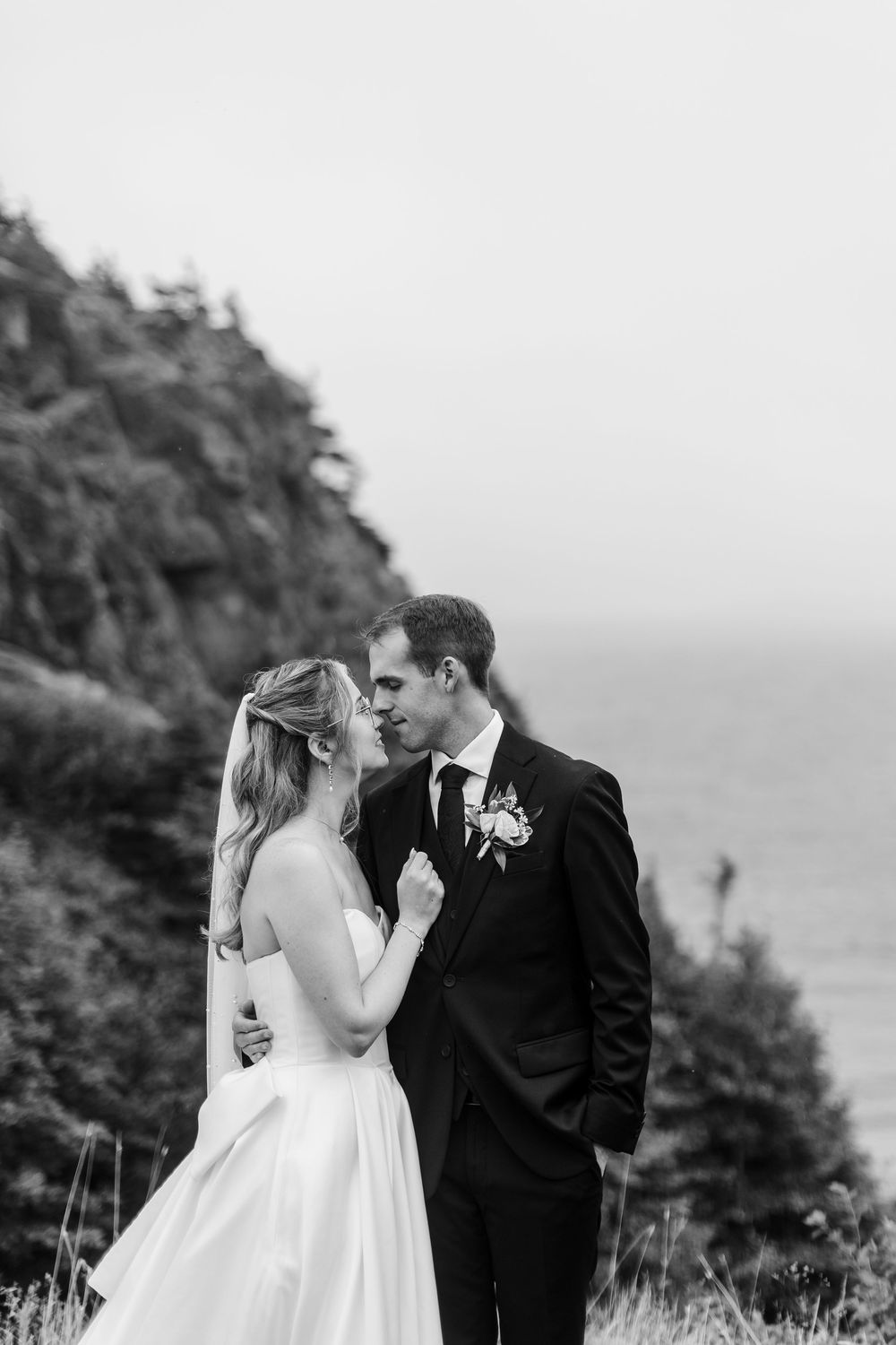 Black and white photo of a bride and groom sharing a romantic moment on a cliffside overlooking the ocean.