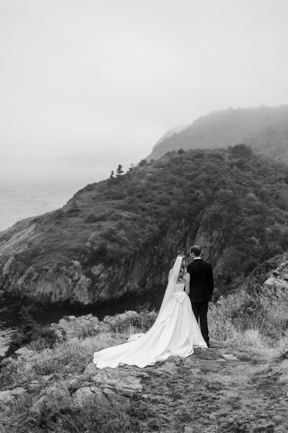 Black and white photo of a bride and groom standing on a rocky cliff overlooking a misty ocean and lush hillside.