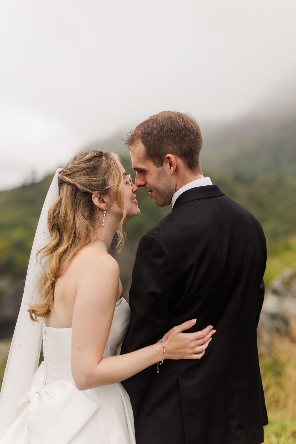 Smiling bride and groom in wedding attire share a romantic moment outdoors with misty mountains behind them.
