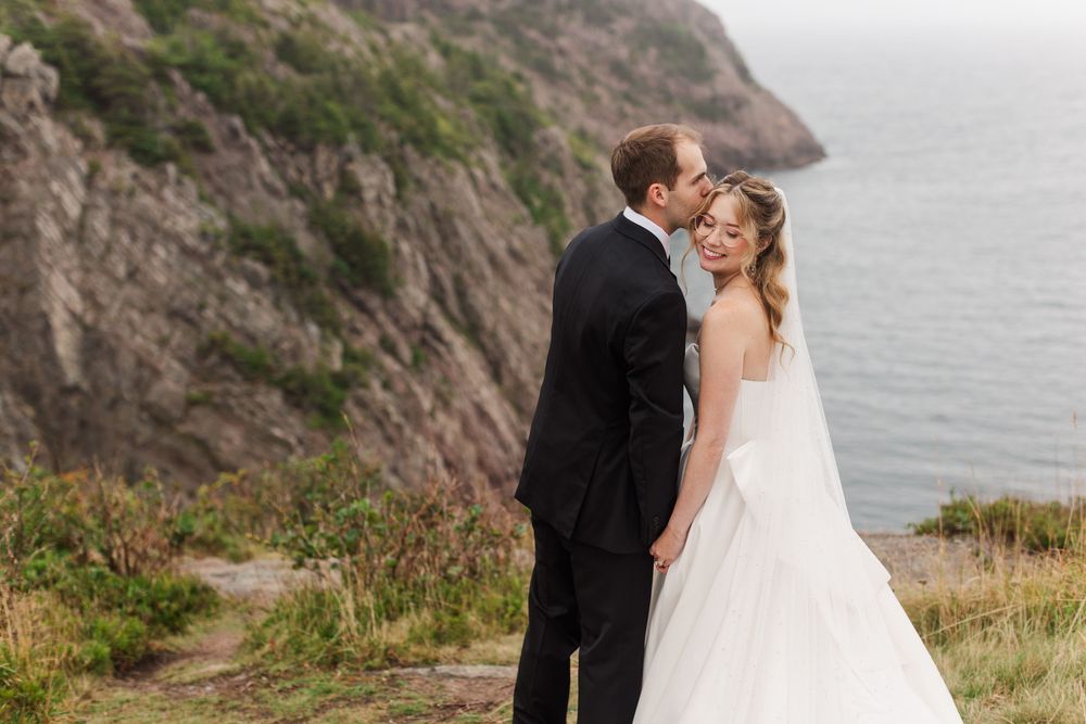 Bride and groom share a romantic kiss on a coastal cliff, with ocean and rocky landscape in the background.