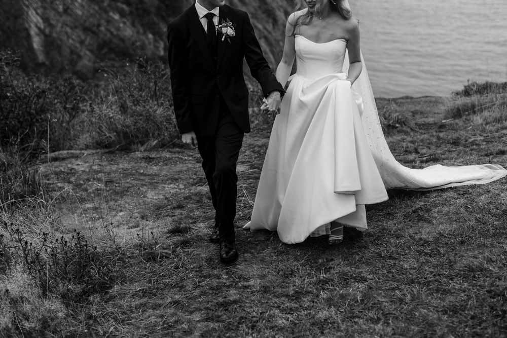 Black and white photo of a bride and groom holding hands walking along a coastal cliff path on their wedding day.