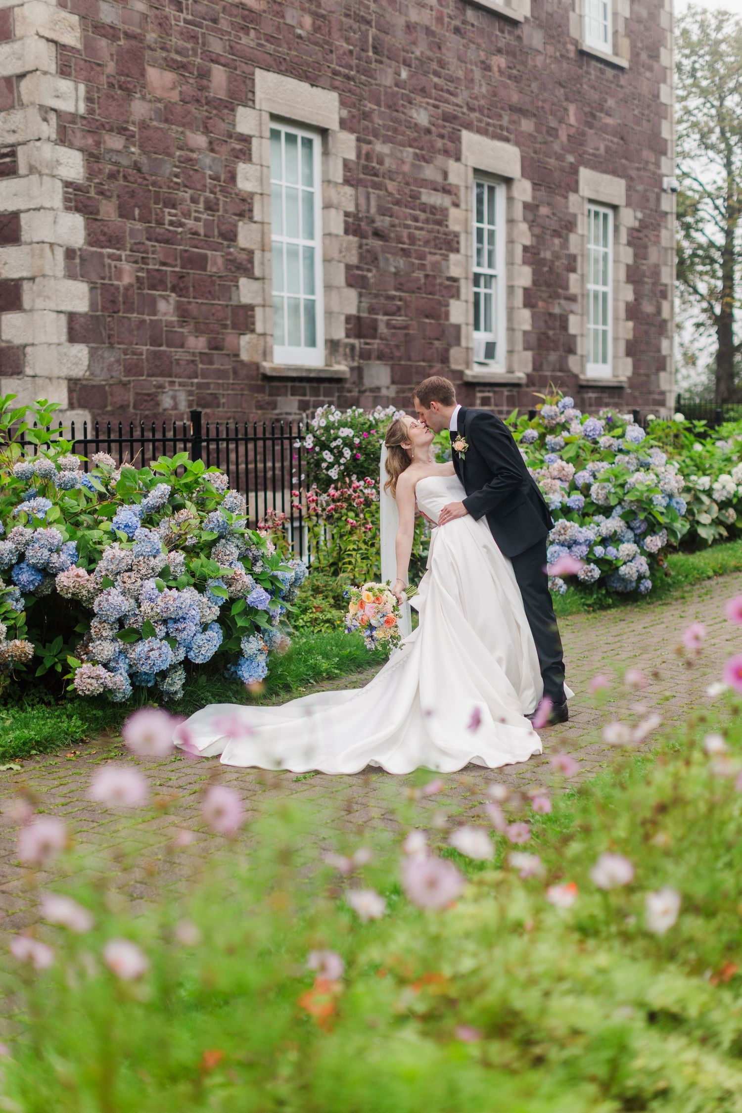 Bride and groom share a kiss in a lush garden with blooming hydrangeas beside a historic stone building.