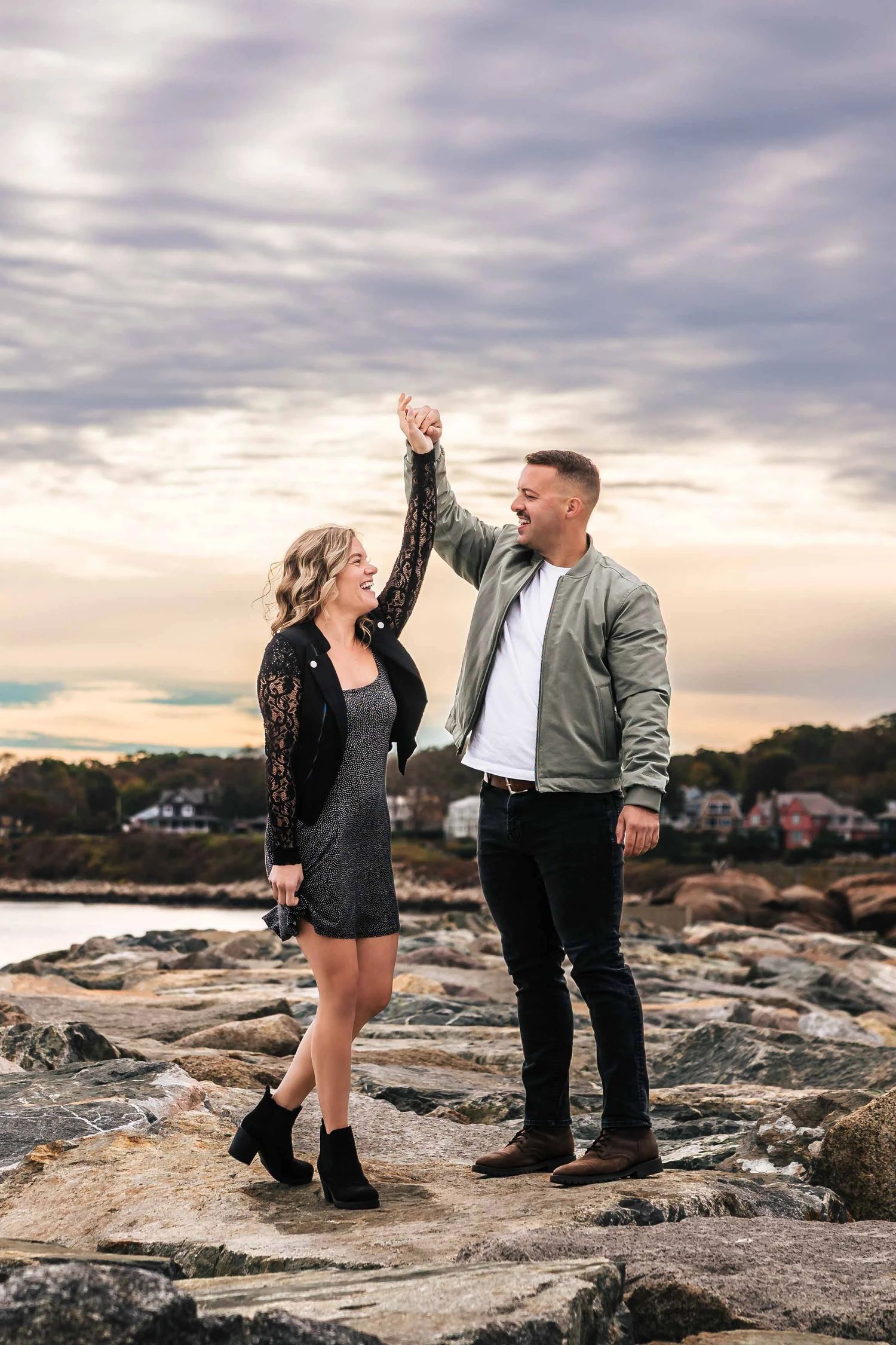 Couple standing together on granite breaker at dawn, woman's hair blowing in wind. Engagement at Rockport Harbor, Rockport, MA