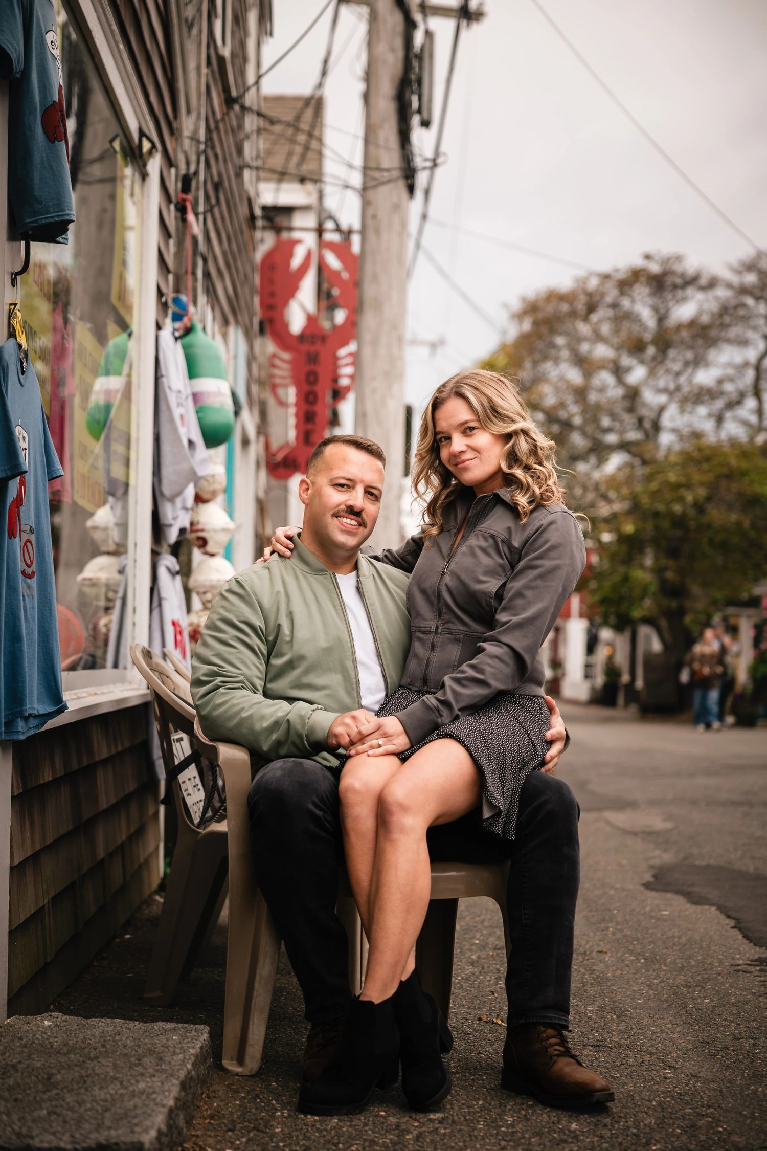 A couple sits together on a bench against a colorful graffitied wall, smiling and holding hands. Engagement at Rockport Harbor, Rockport, MA