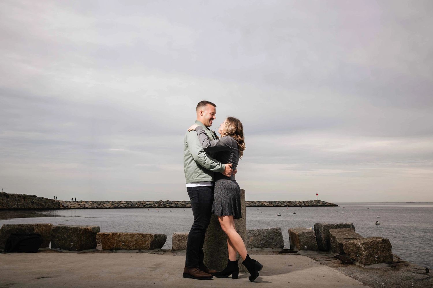 Man lifting woman in his arms while standing on a rocky pier, both smiling at each other. Engagement at Rockport Harbor, Rockport, MA