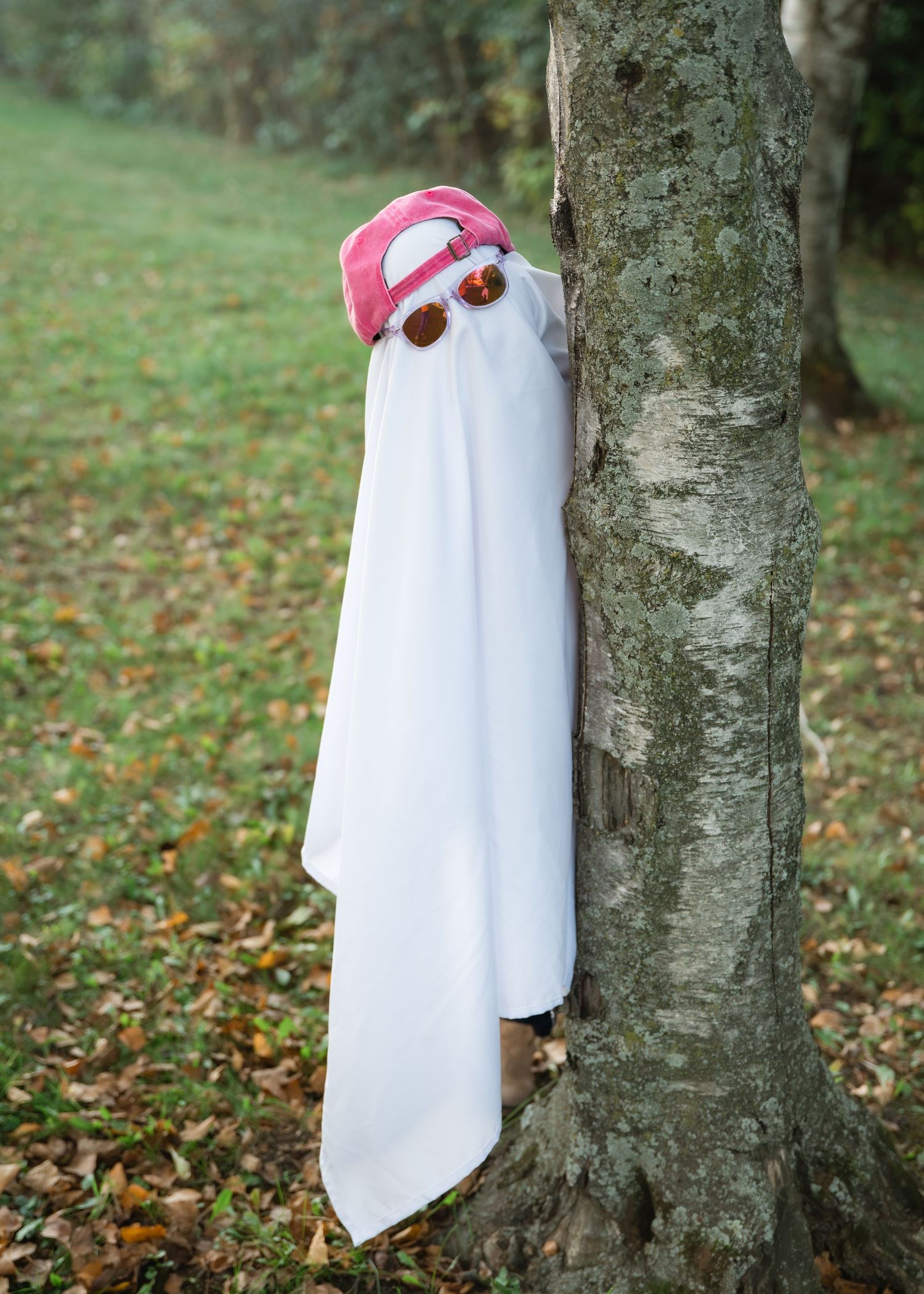 Person in a white sheet ghost costume with a pink cap and round sunglasses peeks from behind a tree trunk on a leaf-covered lawn.