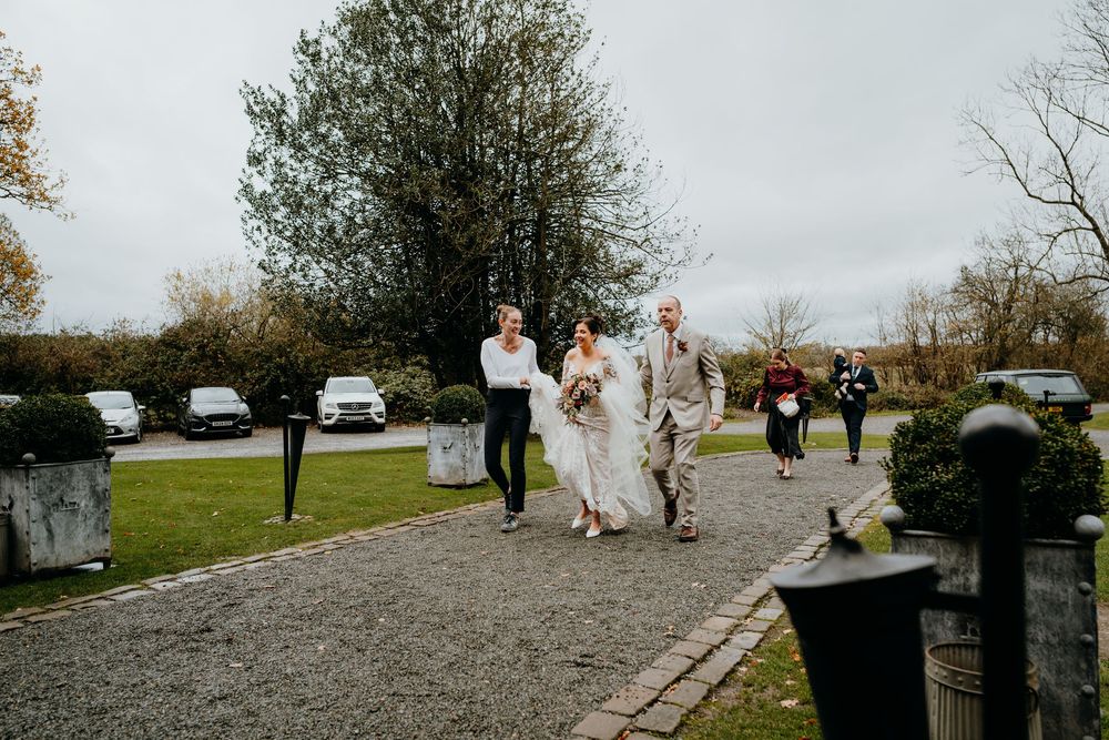 Bride in lace gown walking with escorts along gravel path at outdoor wedding venue in autumn.