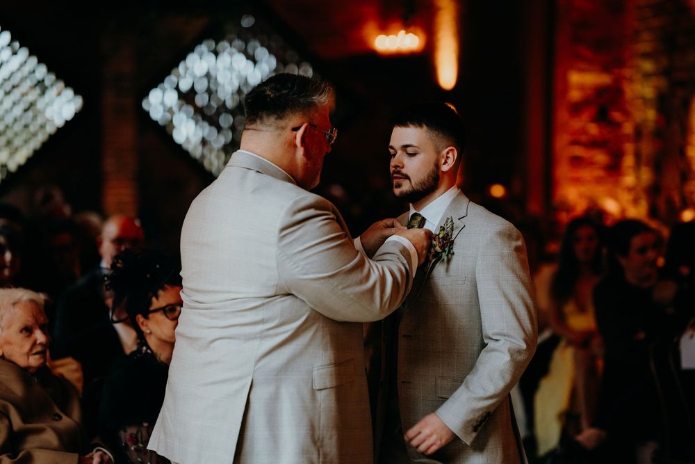 Groom adjusting boutonniere of another groom in light suit during wedding ceremony before guests.