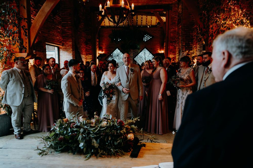 Bride and groom exchanging vows at rustic barn wedding ceremony surrounded by guests and floral decor.