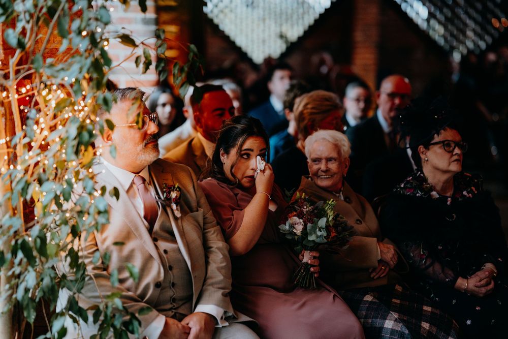 Emotional wedding guests including elderly woman and tearful bridesmaid watching ceremony in warmly lit venue.