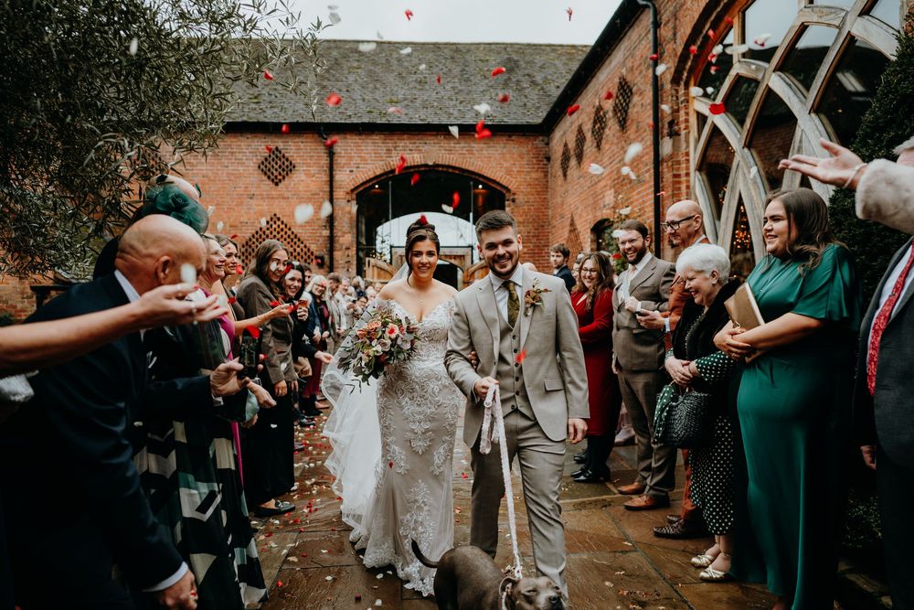 Bride and groom walk through confetti shower with wedding guests cheering in rustic brick courtyard venue.
