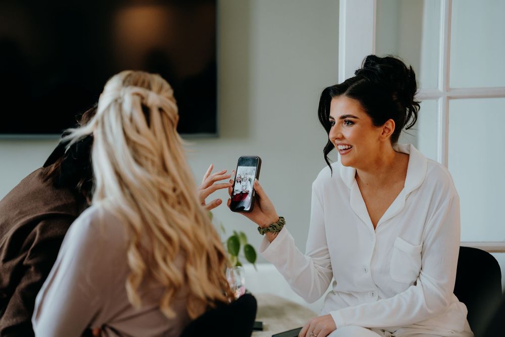 Smiling bride showing phone photo to friend during wedding morning preparations.