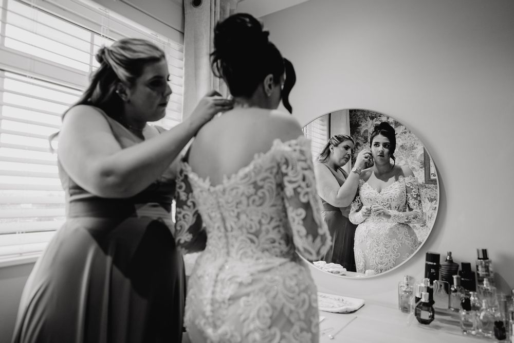 Black and white photo of bridesmaid helping bride into lace wedding dress.