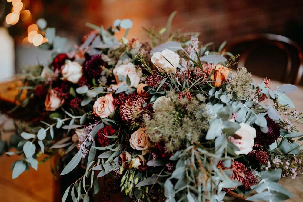 Lush autumn wedding bouquet with burgundy, blush, and peach roses, eucalyptus, and wildflowers on a wooden table.