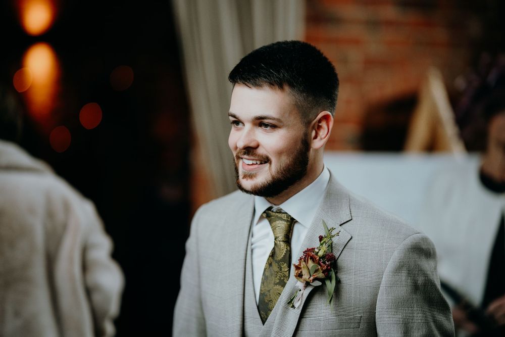 Smiling groom in light grey suit with floral boutonniere during wedding ceremony in warmly lit venue.