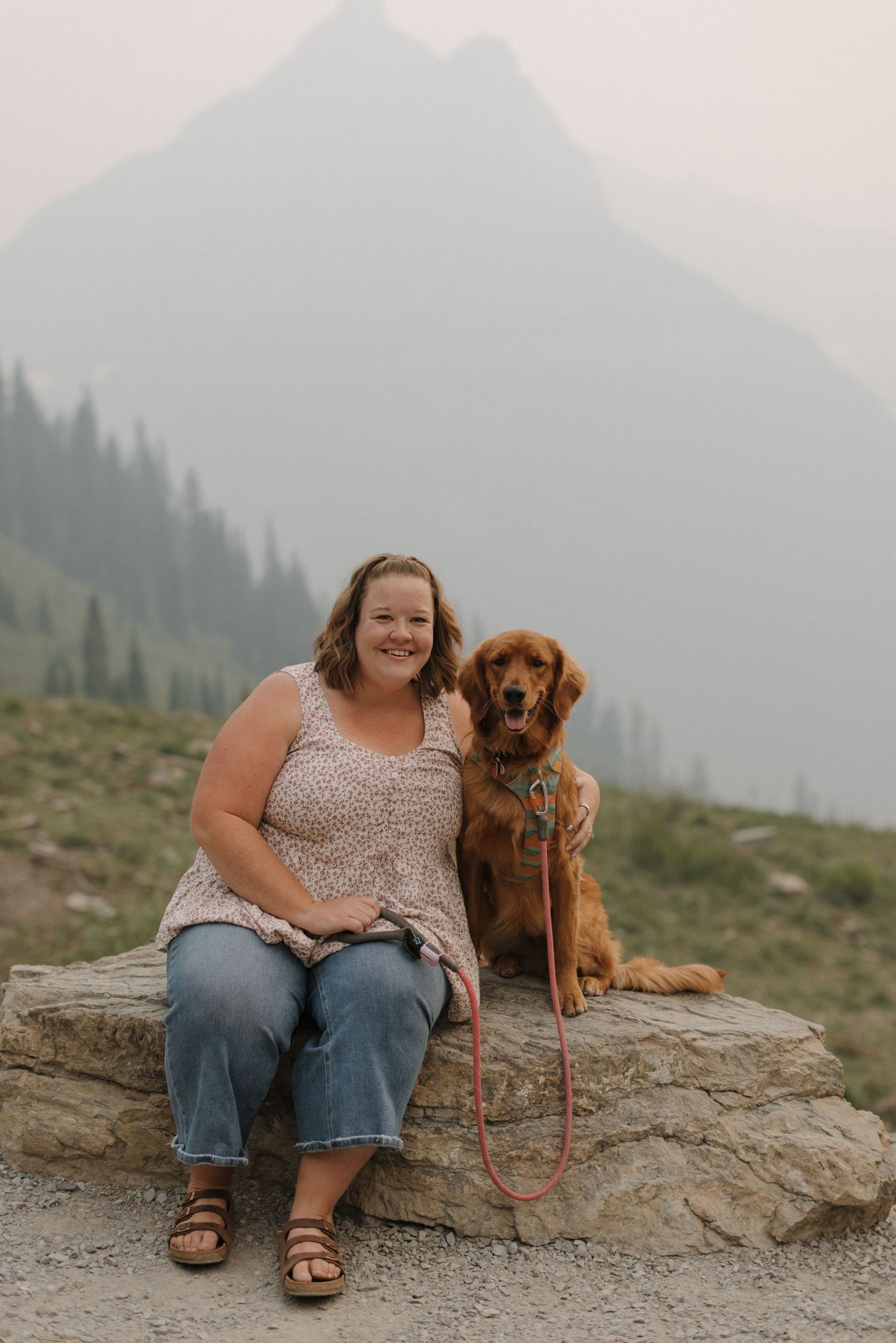 Woman and golden retriever dog sitting on a rock with misty mountain landscape in the background.