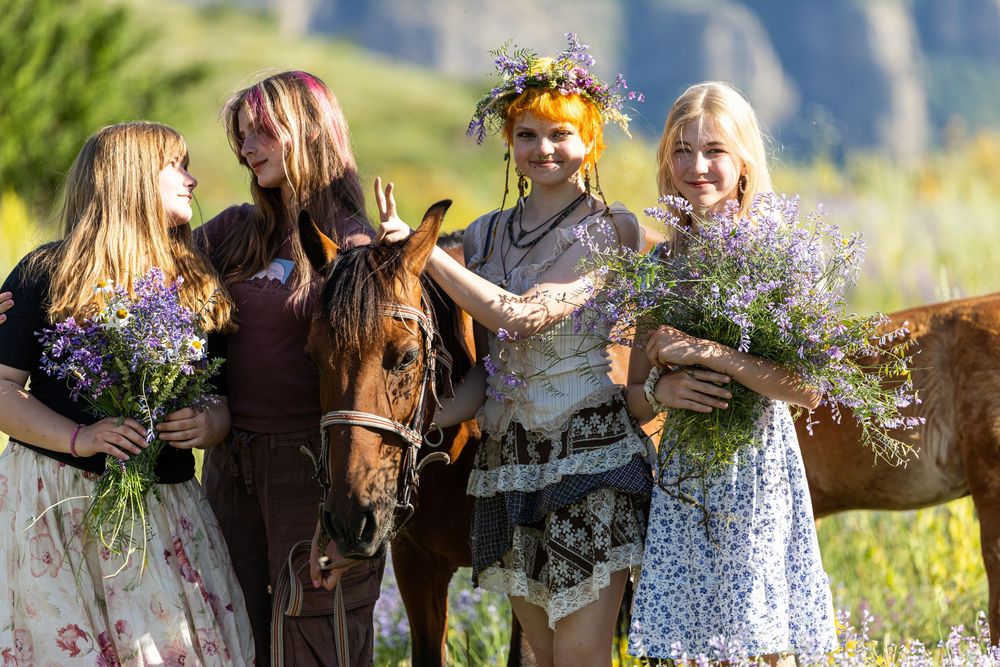 Four young women in boho dresses posing with a brown horse holding wildflower bouquets outdoors.
