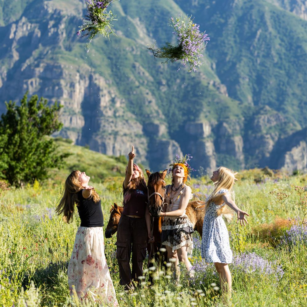 Group of young women tossing wildflower bouquets in the air in a scenic mountain meadow.