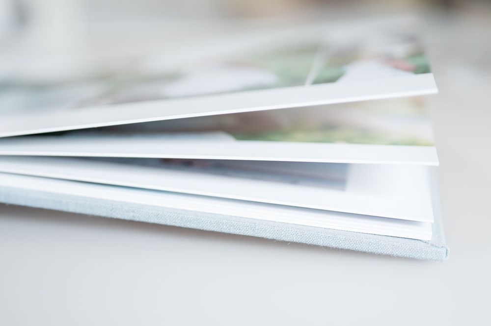 Close-up side view of an open hardcover photo book with thick white pages and a light blue linen cover resting on a white surface.