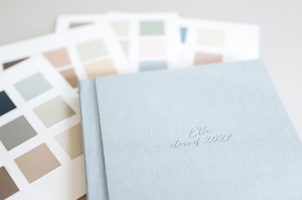 Light blue linen-bound photo album embossed with Elle Class of 2021 resting on neutral color swatch cards in soft beige and gray tones.