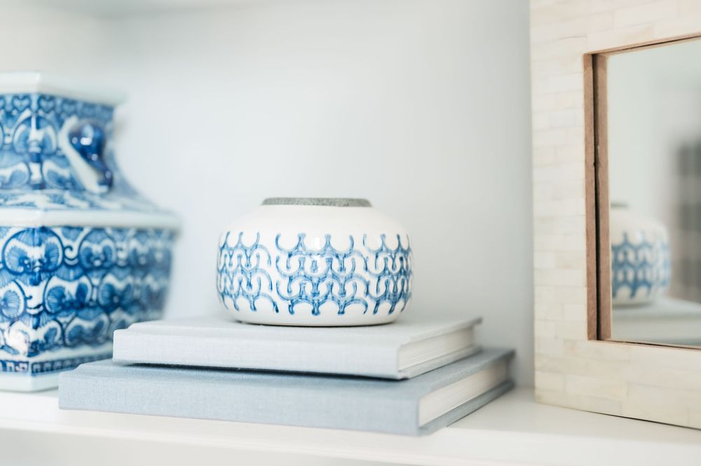 Blue and white patterned ceramic vase resting on stacked books on a white shelf beside a larger porcelain jar and a bone inlay mirror.