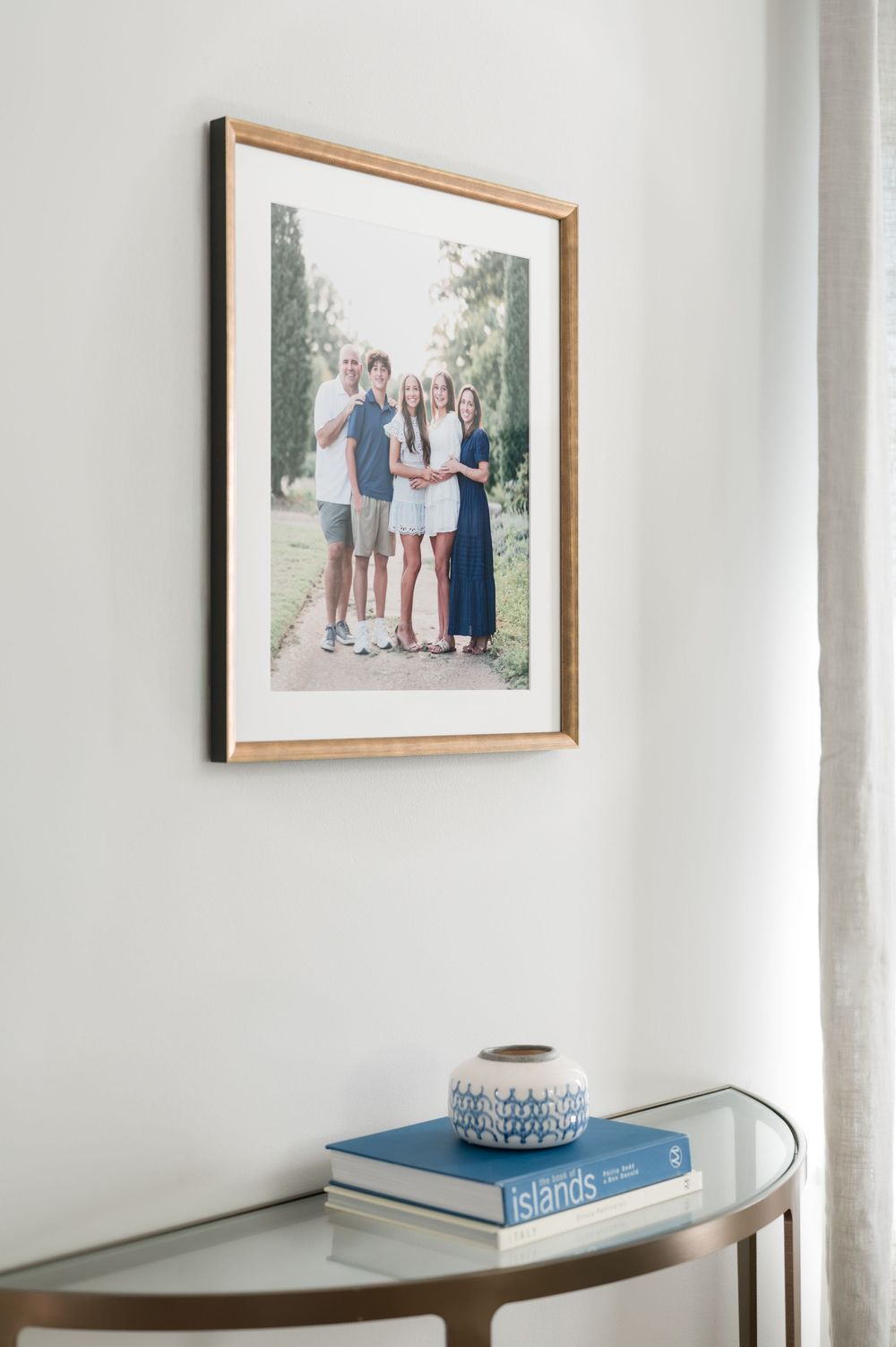 Framed family portrait of five people standing outdoors hangs above a glass console table with a blue Islands book and a decorative vase.