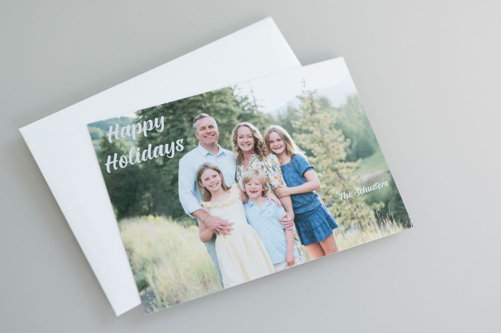 Happy Holidays photo card showing a smiling family of five posing outdoors in a grassy field with trees, resting on a white envelope.