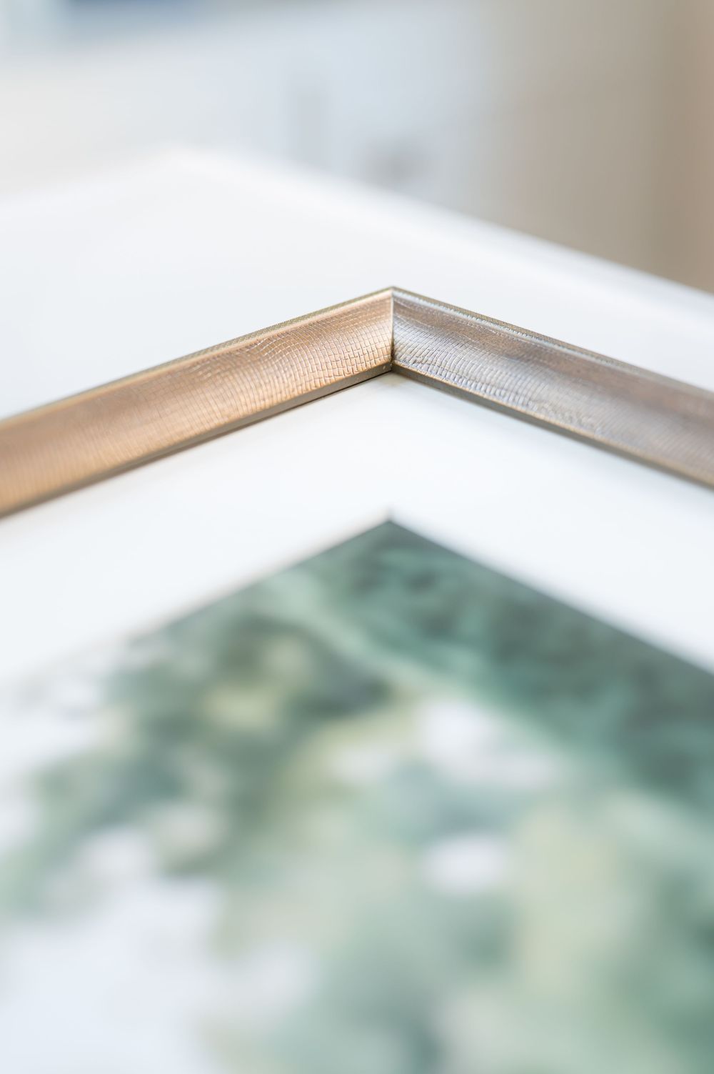 Close-up of a brushed gold picture frame corner with a white mat and a blurred green watercolor print inside.