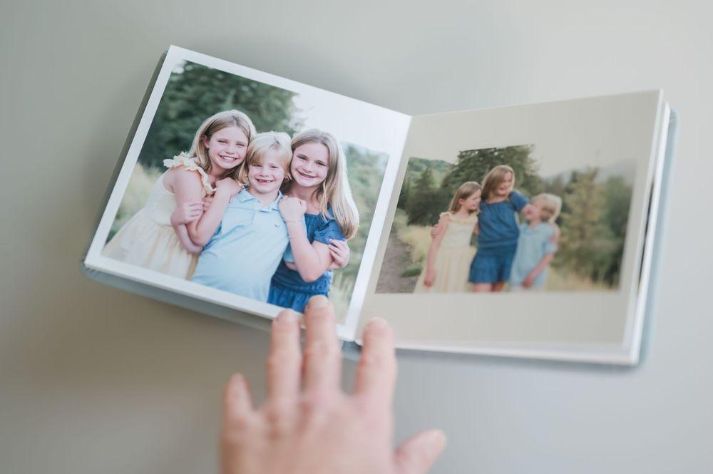 Hand resting beside an open photo album showing three smiling girls hugging outdoors and a family portrait among green trees.