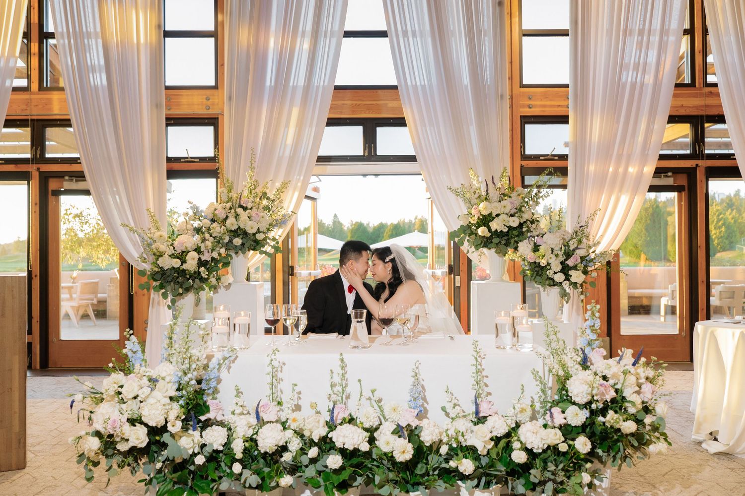 Bride and groom share a kiss at elegant white floral sweetheart table with draped fabric backdrop at wedding reception.