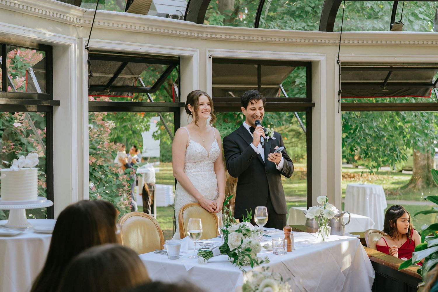 Bride and groom share a joyful moment during wedding reception toast inside elegant glass conservatory venue.