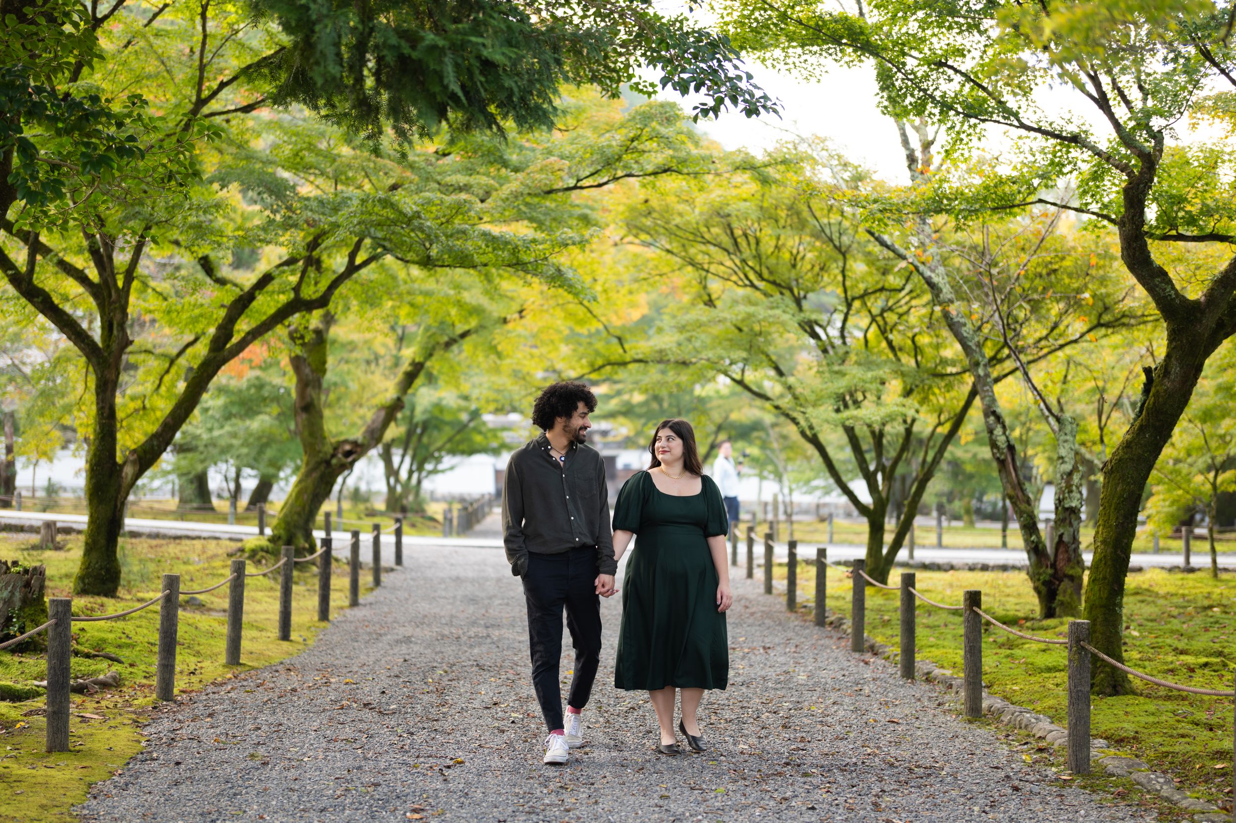 Couple walking hand in hand along a tree-lined gravel path in a lush Japanese garden.