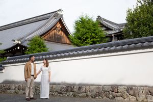 Couple holding hands in front of a traditional Japanese temple wall with tiled rooftops and lush greenery.