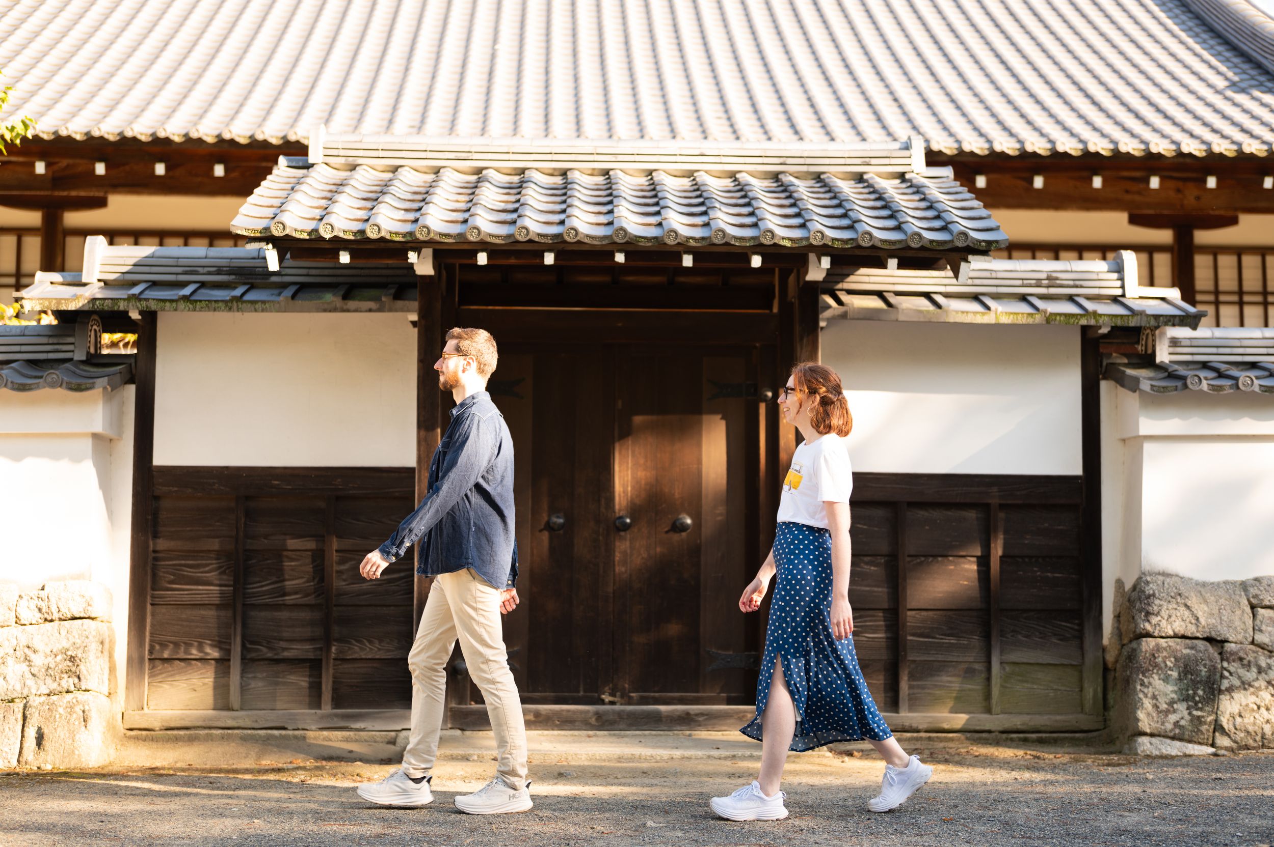 Couple walking past a traditional Japanese building with white walls and dark wooden gates in Kyoto, Japan.
