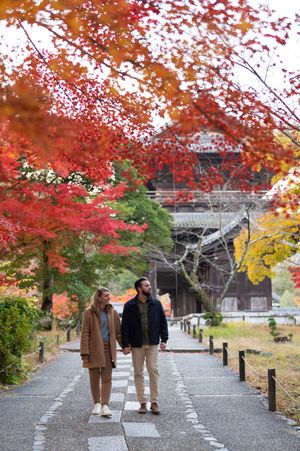Couple holding hands walking on a stone path surrounded by vibrant red autumn maple trees near a Japanese temple.