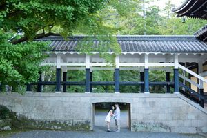 Couple standing under a traditional Japanese temple bridge surrounded by lush green trees.