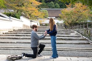 Man proposing to woman on one knee at a scenic Japanese temple with stone steps and autumn trees.