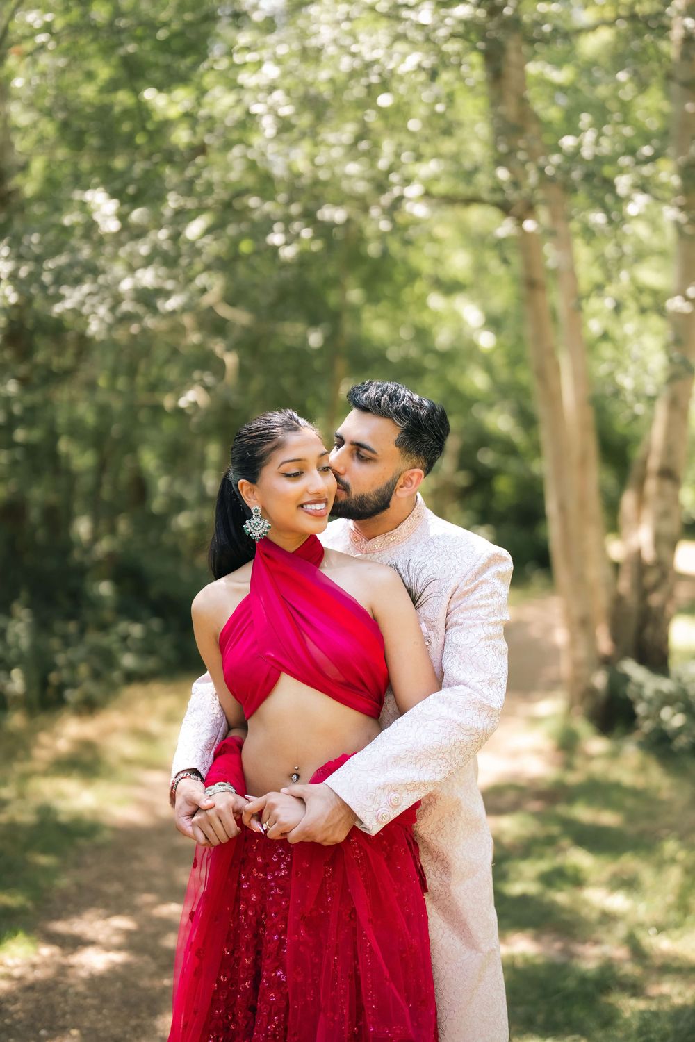 Indian couple in traditional attire embracing outdoors, woman in red lehenga, man in white sherwani, lush green trees.