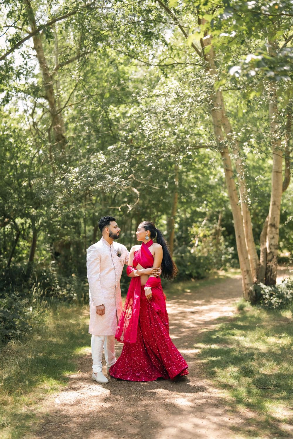 Indian couple in traditional wedding attire stand on a woodland path, the bride in red lehenga and groom in ivory sherwani.