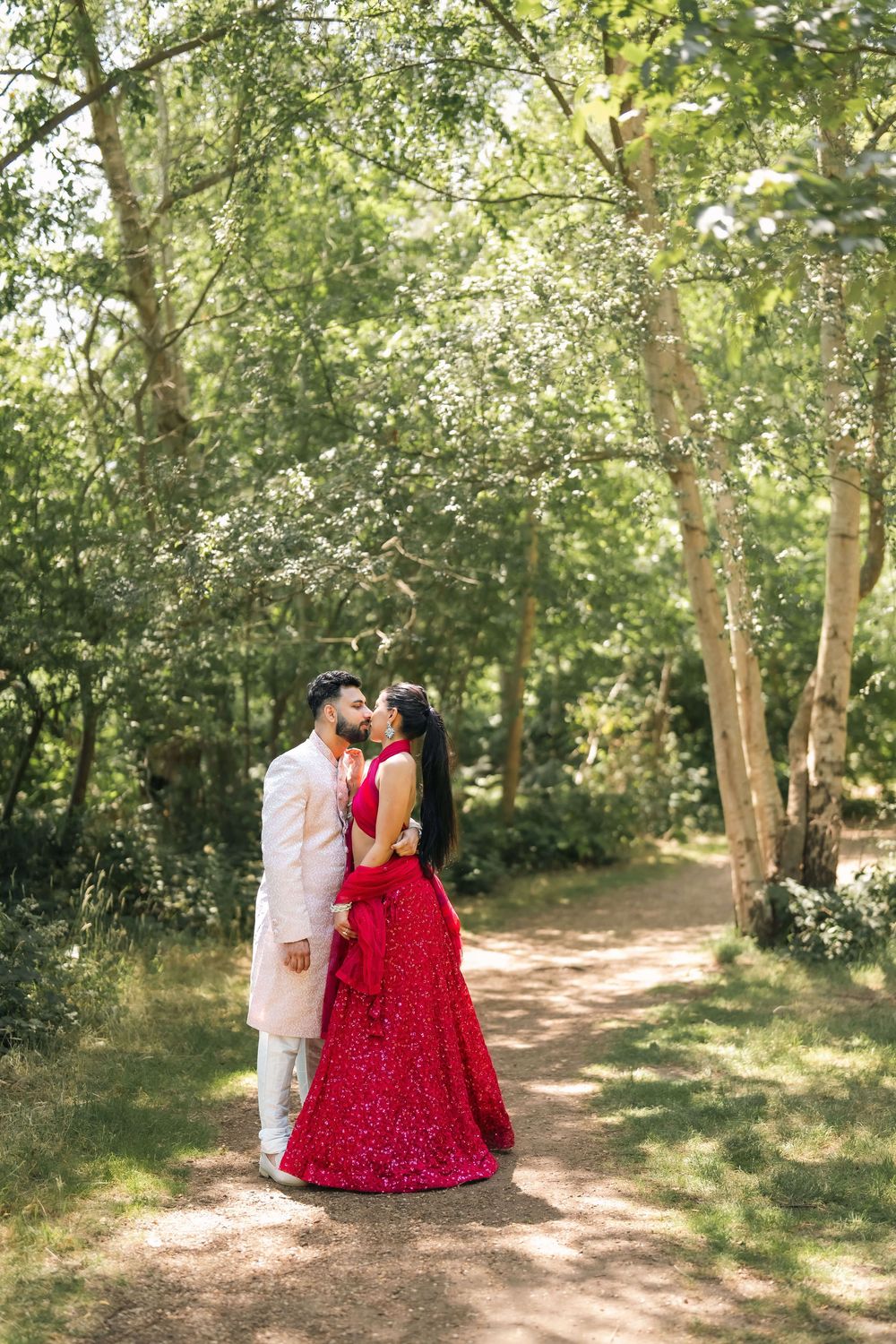 A couple in traditional Indian wedding attire share a romantic moment on a forest path, bride in red lehenga.