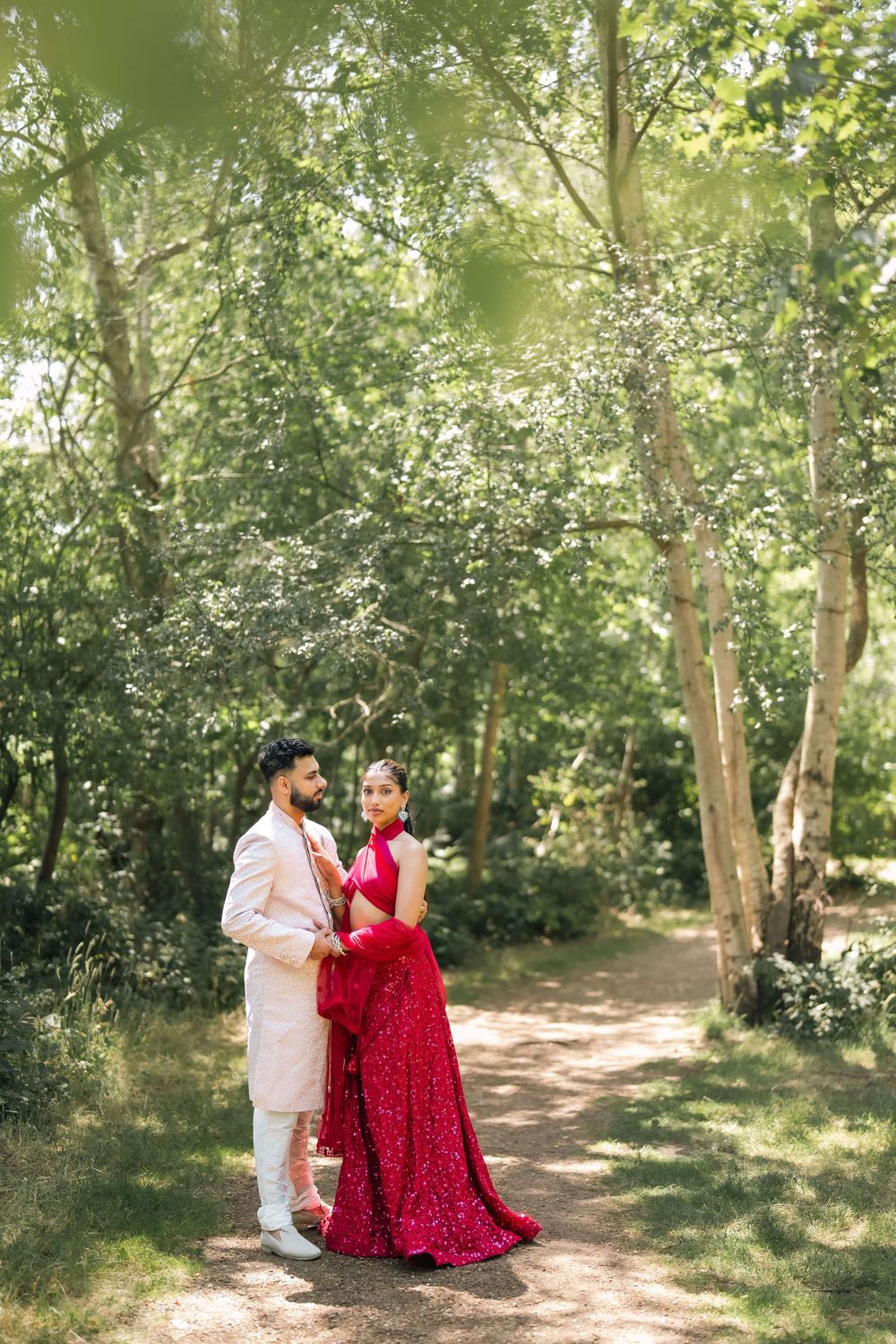 South Asian couple in traditional wedding attire posing on a sunlit forest path, bride in red gown, groom in ivory suit.
