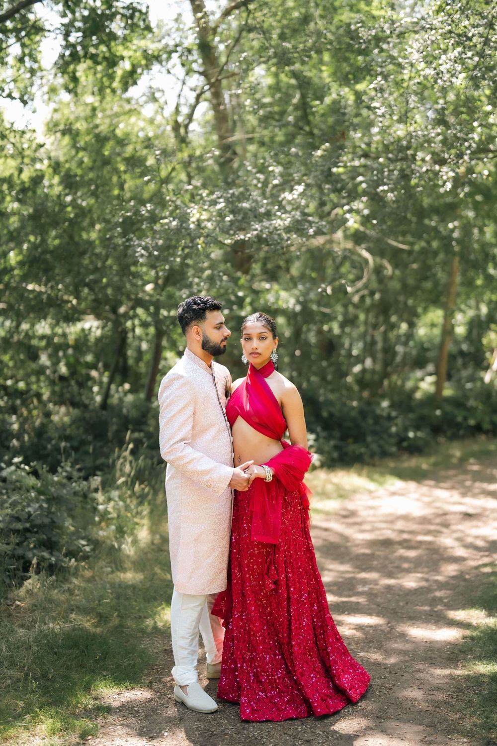 Couple in traditional Indian wedding attire posing in lush green forest, woman in red lehenga, man in cream sherwani.
