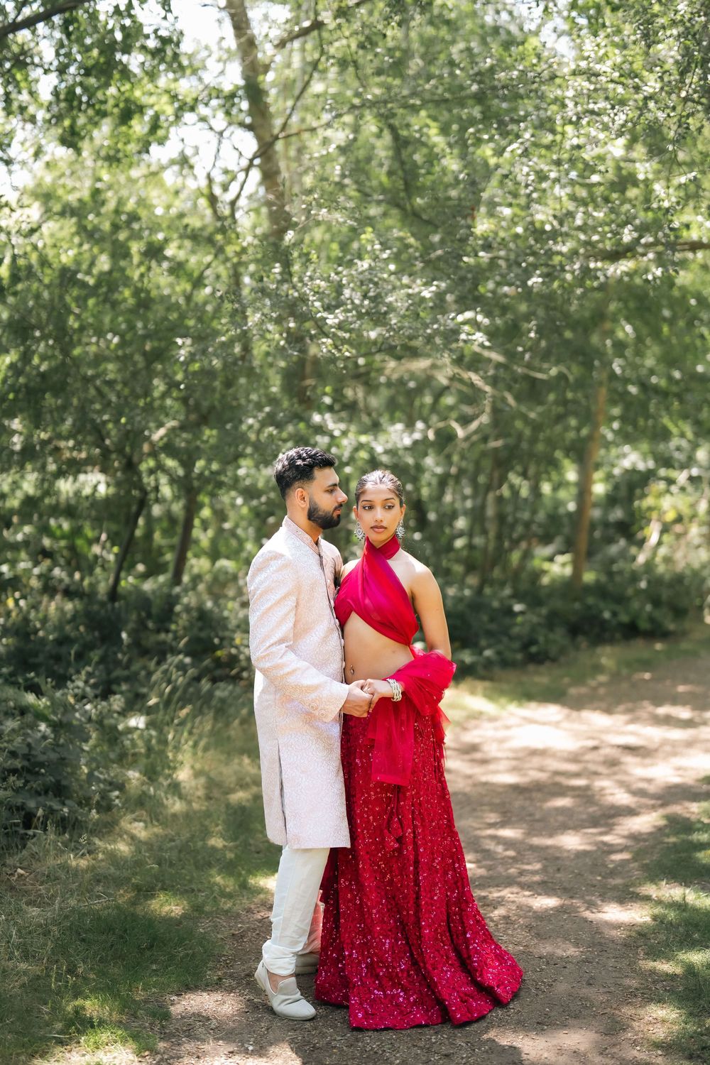 Couple in traditional Indian wedding attire posing on a garden path, woman in red lehenga, man in cream sherwani.