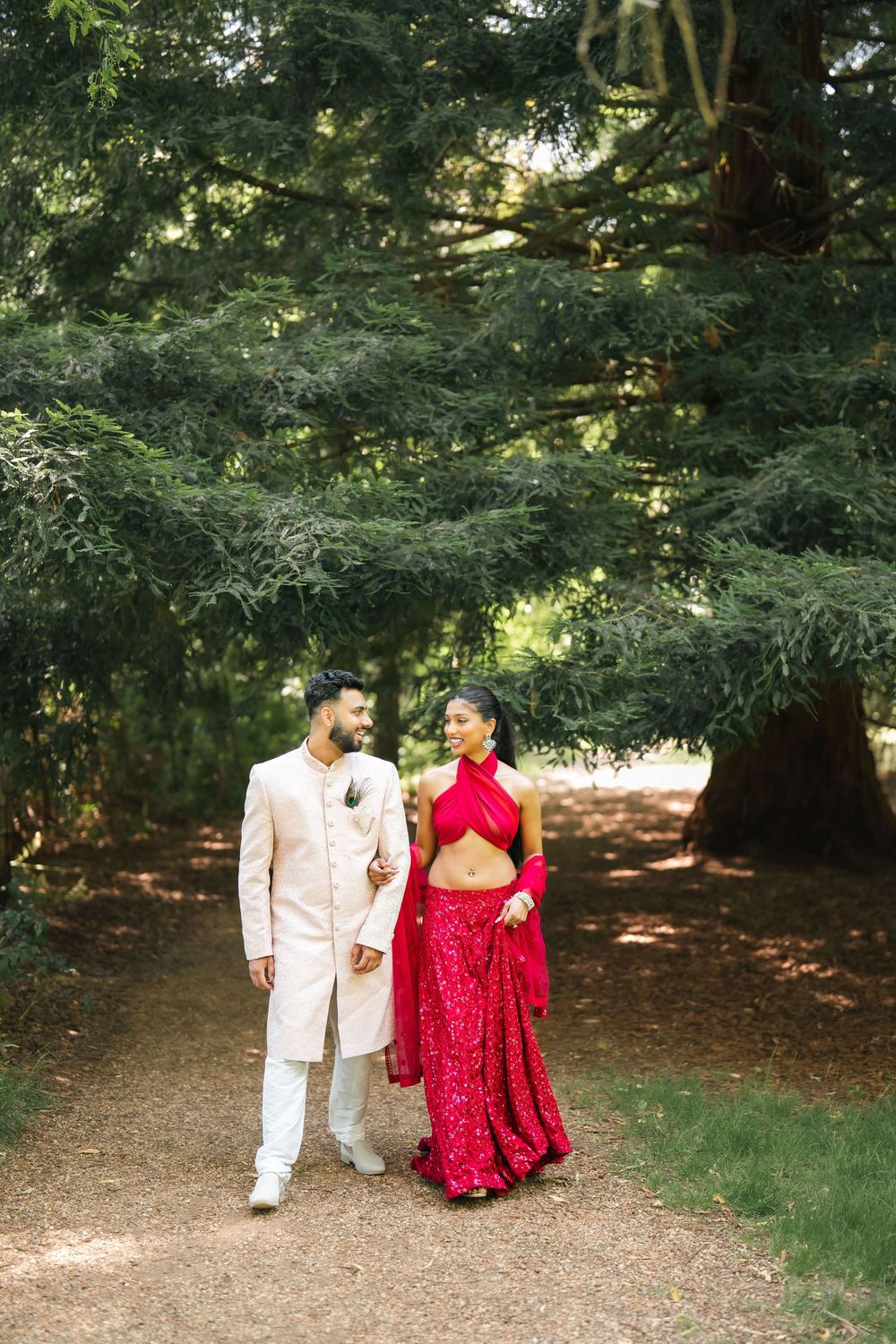 A couple in traditional South Asian wedding attire walks hand in hand along a garden path surrounded by lush green trees.