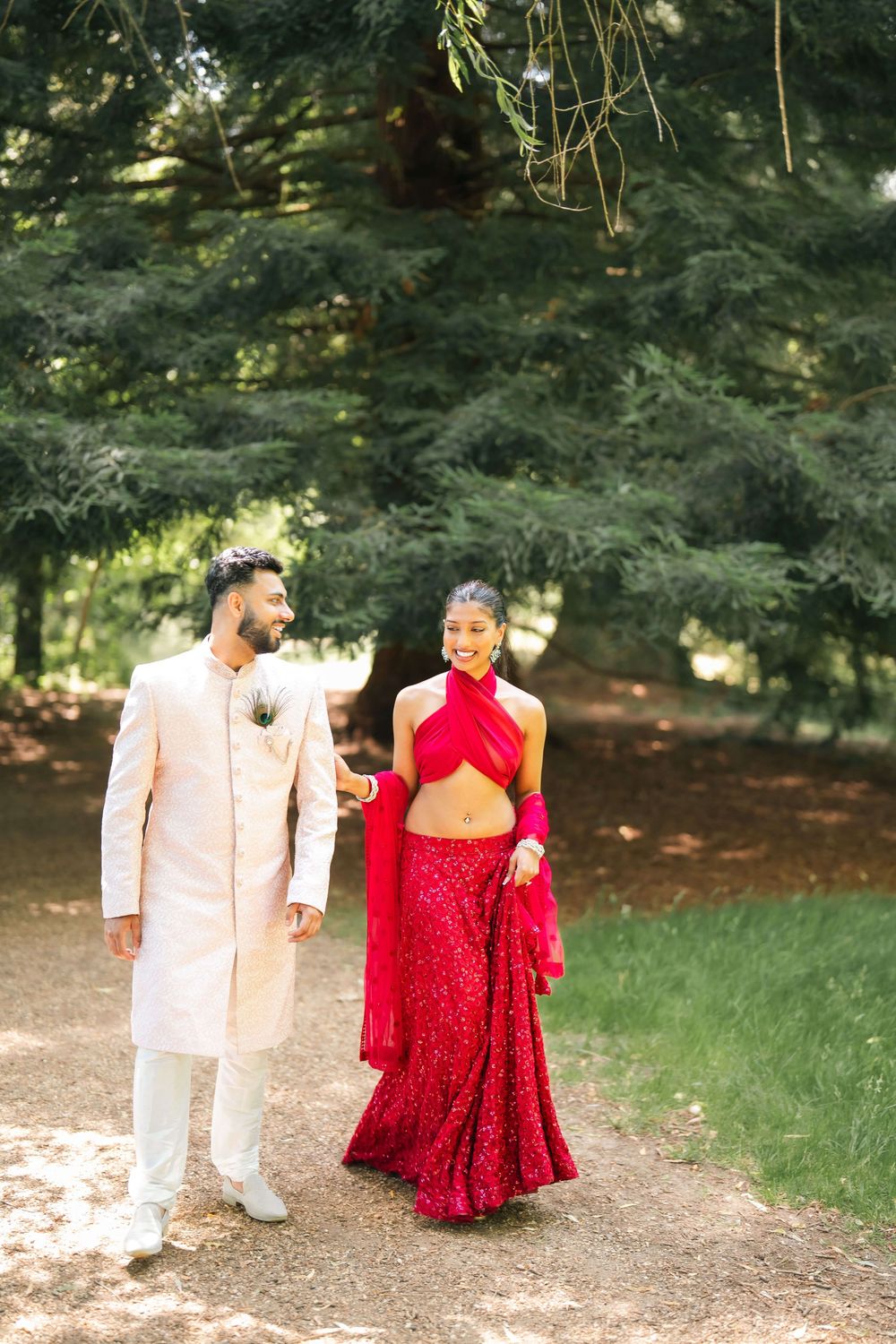 A couple in traditional South Asian wedding attire walks through a lush green park, the bride in red lehenga and groom in cream sherwani.