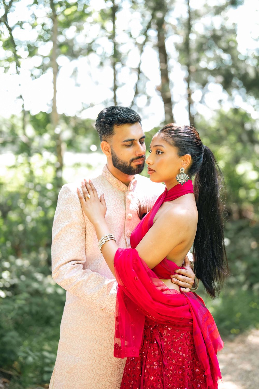 A couple in traditional South Asian attire embrace outdoors, the woman in red and the man in a cream sherwani.