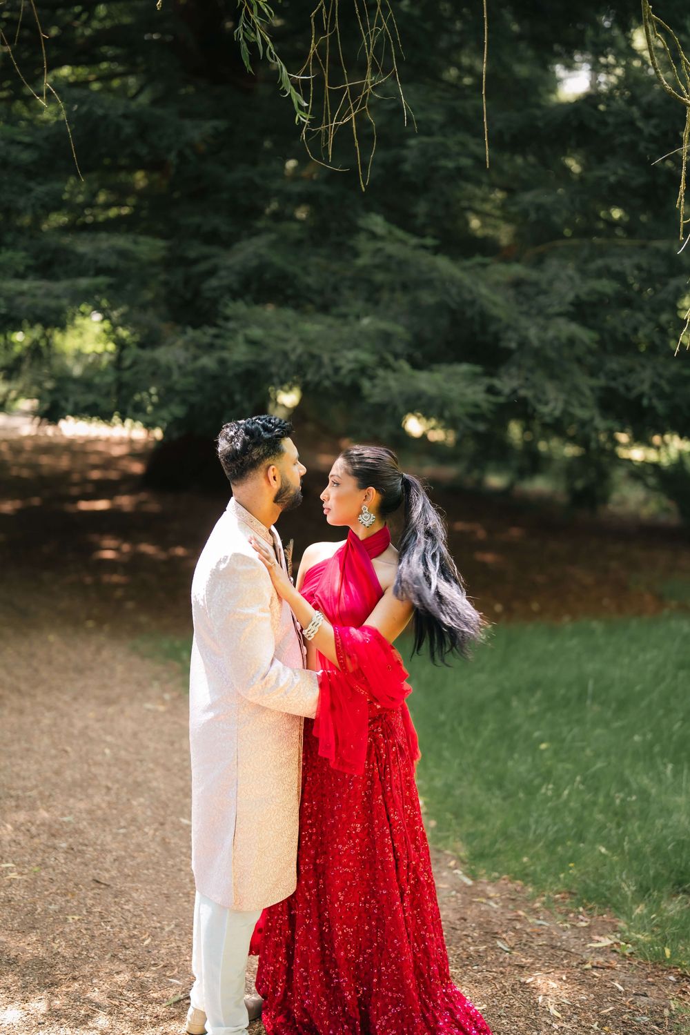A couple in traditional South Asian wedding attire embraces on a sunlit path surrounded by lush green trees.