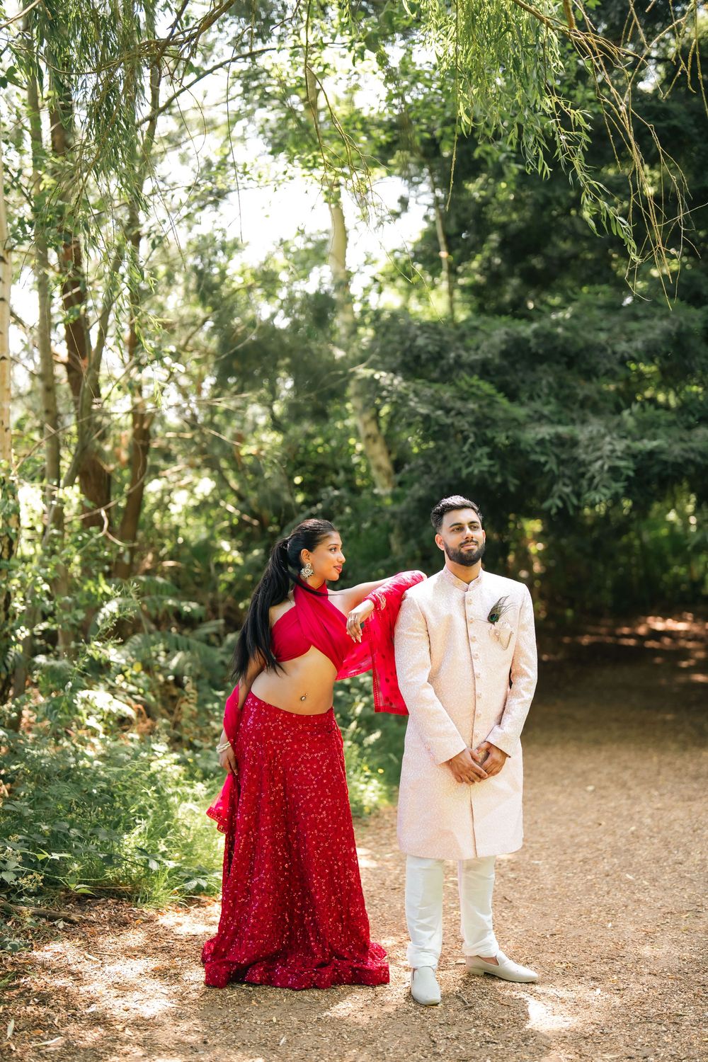 Couple in traditional South Asian attire posing outdoors, woman in red sequin saree and man in cream sherwani.
