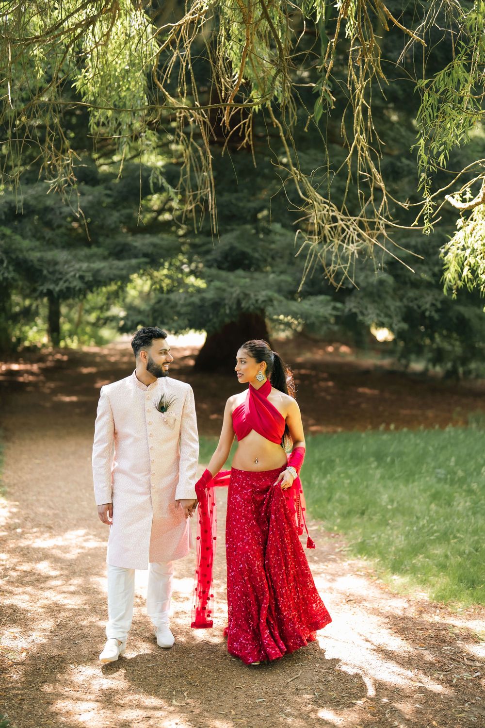 Indian couple in traditional wedding attire walk through a sunlit garden path, groom in cream sherwani, bride in red lehenga.